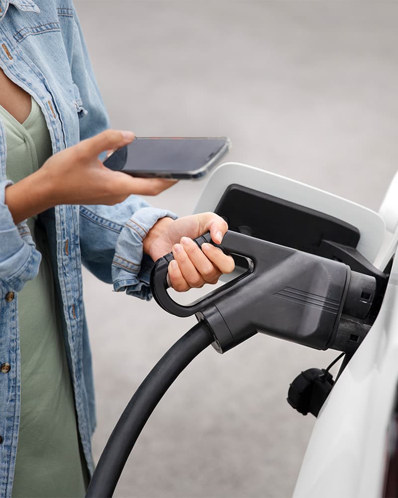Frau hält Smartphone und lädt gleichzeitig ein Elektroauto an einer Schnellladestation